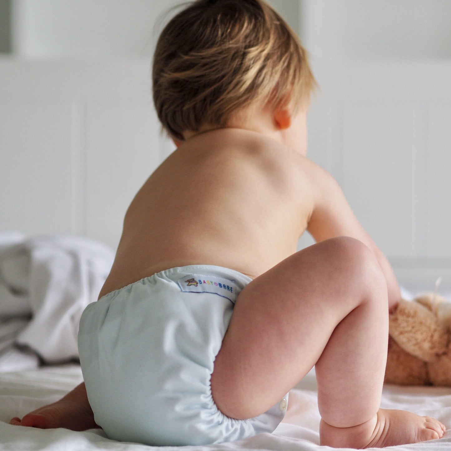 Toddler playing with a teddy bear facing away and wearing a blue nappy. 