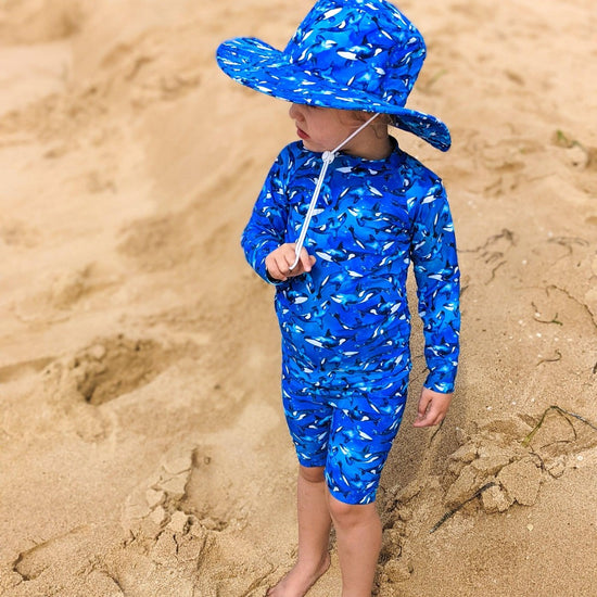Young child standing in the sand wearing long sleeve swimsuit and hat